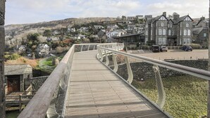Un pont piétonnier en bois, doté d’une rambarde métallique, mène à un parking où sont garées des voitures et offre une vue sur un coteau parsemé de maisons.