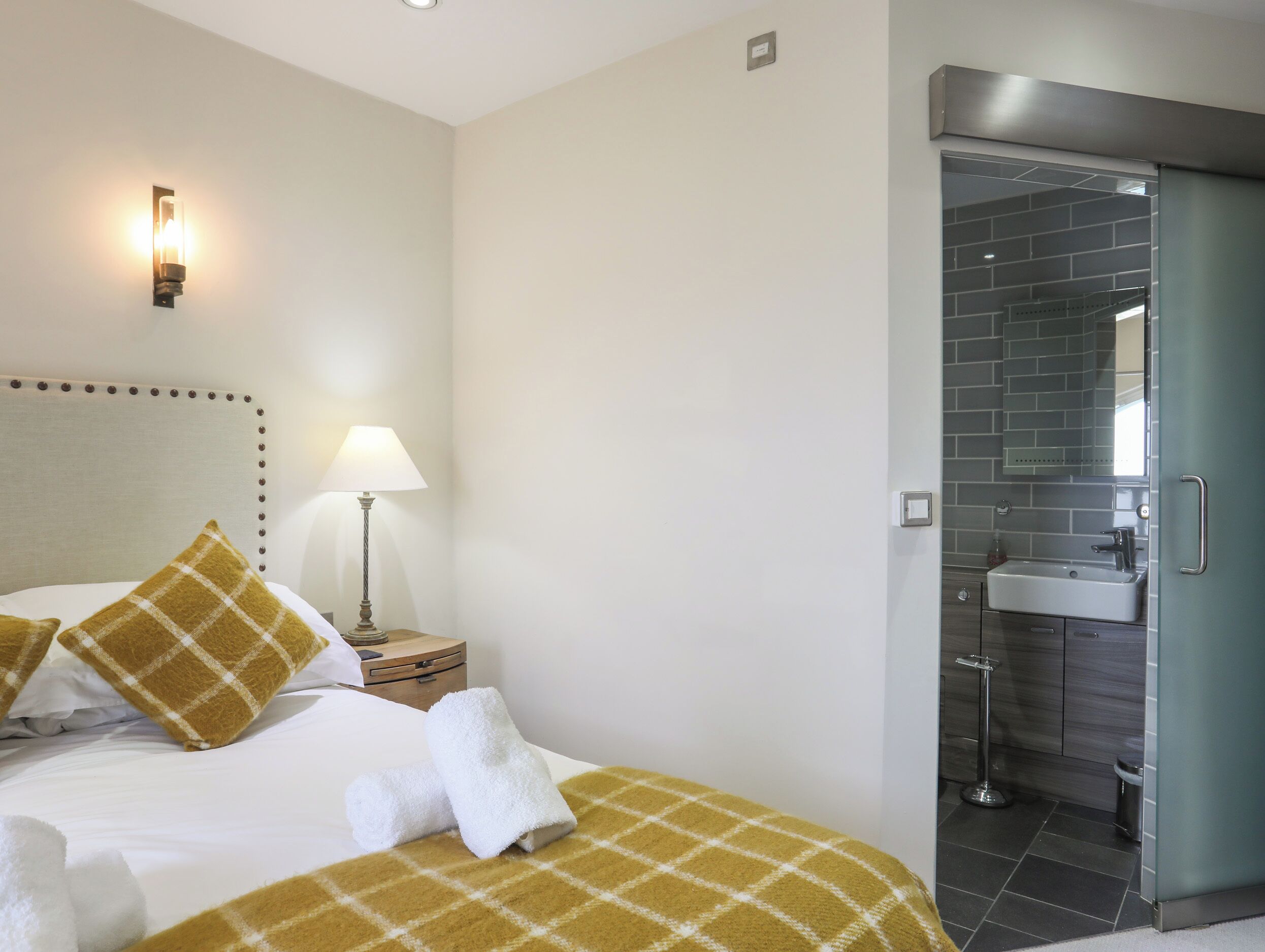 A hotel room with a bed, two pillows, a bedside lamp, a nightstand, and a bathroom with a sink and mirror.