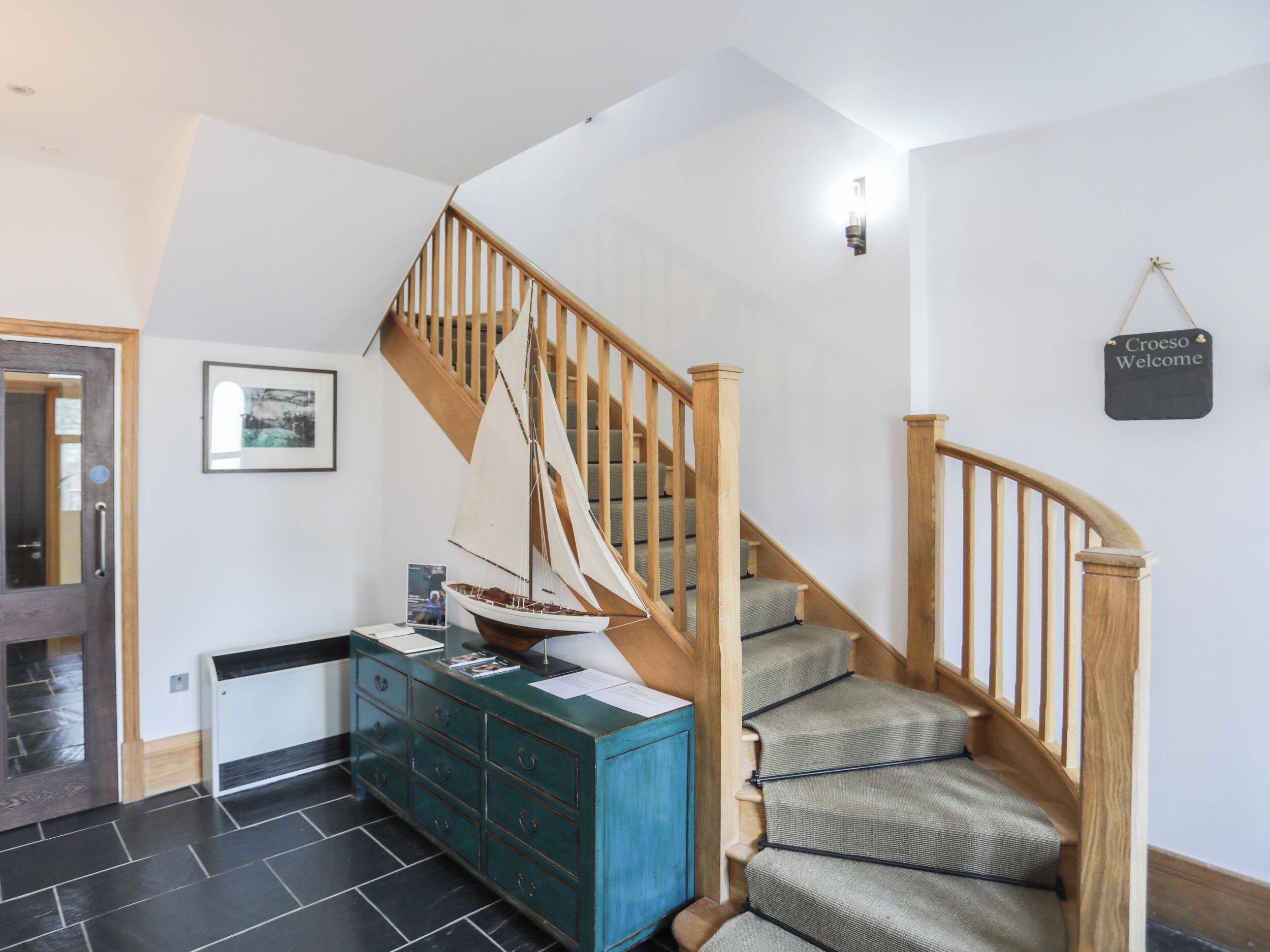 A staircase with wooden railings and carpeted steps, a blue cabinet with drawers, a framed picture on the wall, and a 'Croeso Welcome' sign.