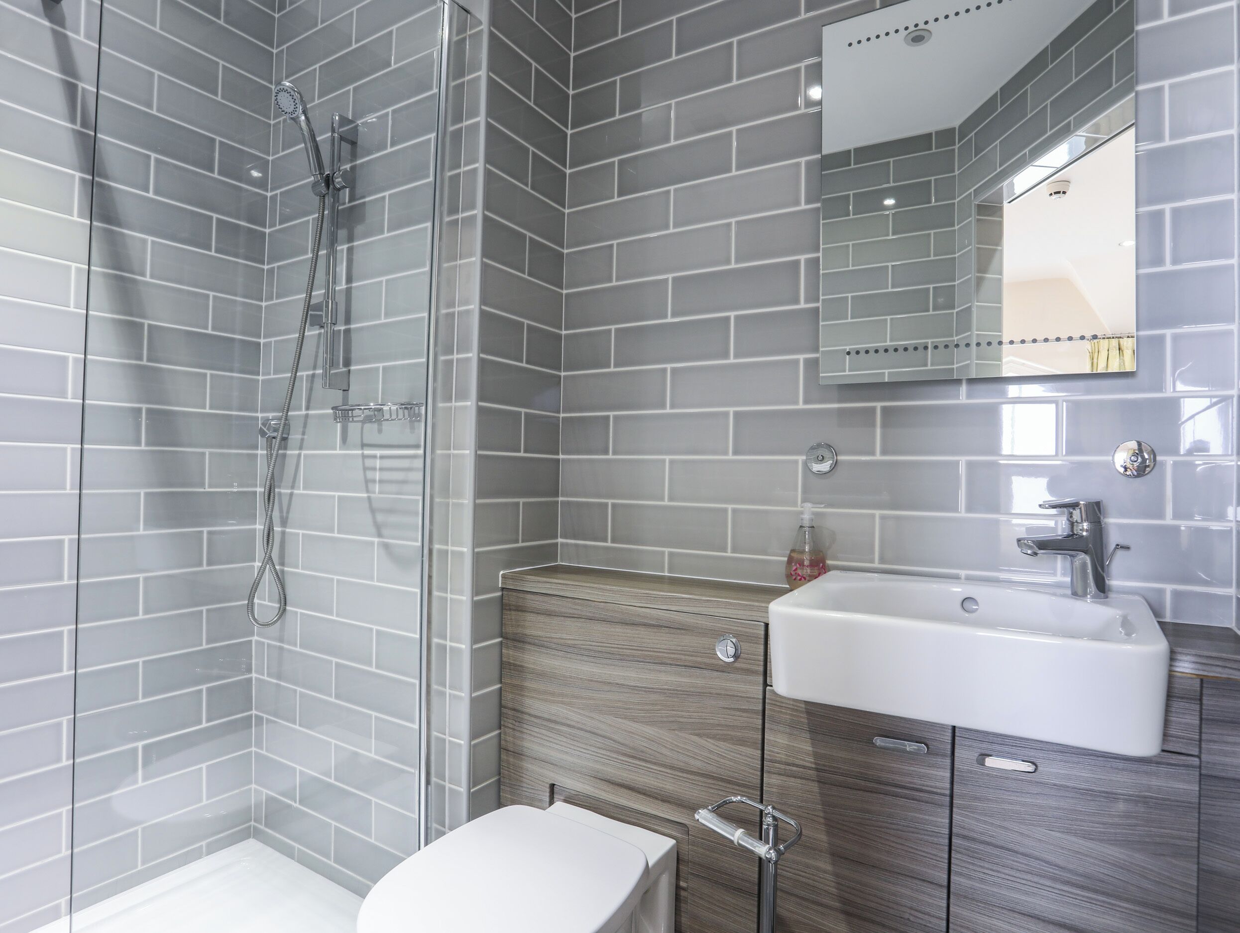 Un baño moderno con paredes de azulejos grises, cabina de ducha con mampara de vidrio, lavabo blanco y tocador de madera.