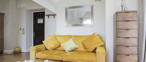 Uma sala de estar com um sofá amarelo, uma mesa de centro de vidro com conjunto de chá, um armário de madeira e um espelho na parede.