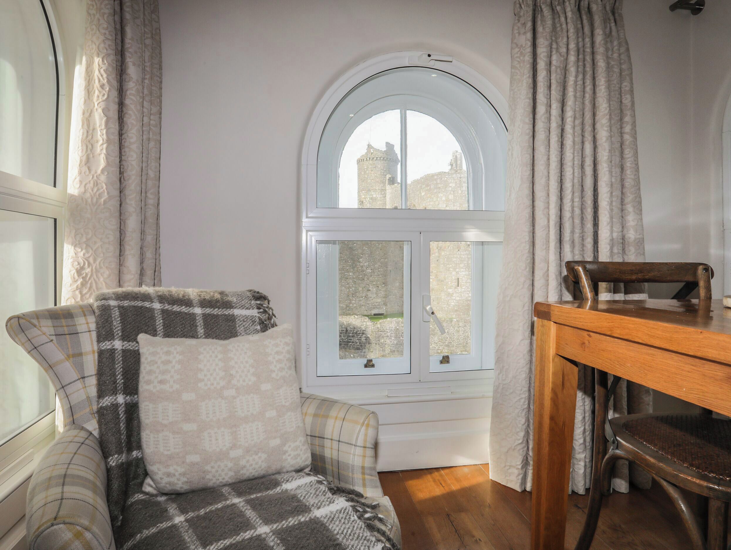 A cozy room with a checkered armchair, a wooden table, and a window with a view of a castle.