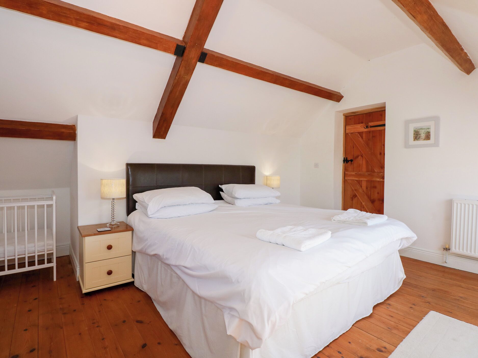 A bedroom with a large bed, wooden beams on the ceiling, a white crib, and a wooden door.