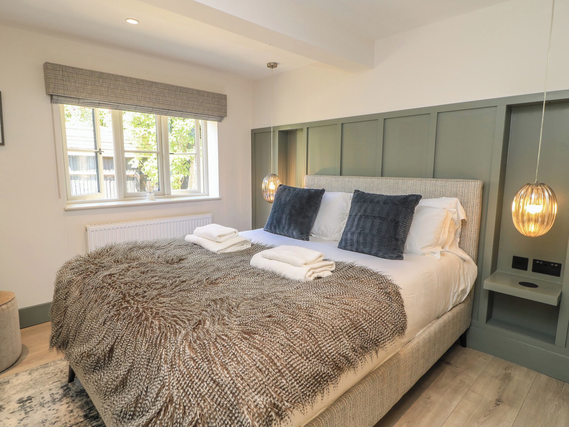 A bedroom with a large bed, a window with blinds, a radiator, and a bedside table with a lamp.