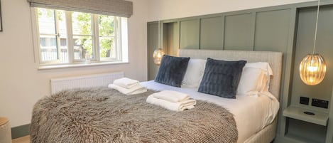 A bedroom with a large bed, a window with blinds, a radiator, and a bedside table with a lamp.