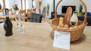 A dining table with a bottle of wine, glasses, a wicker basket with bread, eggs, and a milk bottle, and a welcome card. - The Clockhouse (Montgomery)
