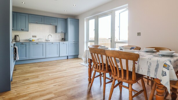 A modern kitchen with blue cabinets, a wooden dining table with chairs, and a large window providing natural light. - 4 Llain yr Eglwys (Newport)