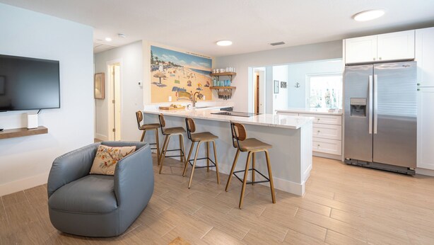 A modern kitchen with a central island, bar stools, a flat-screen TV, and a refrigerator. - 3BR/2BA Luke's Landing Island Home - Jensen Beach Retreat (Jensen Beach)