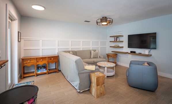Una sala de estar con sofá, un sillón azul, una mesa de centro, televisor y estanterías de madera con libros y adornos.