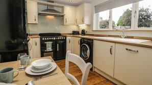 A compact kitchen with a stove, oven, washing machine, and a dining area with a table set for two. - 1 Springfort Cottages (Penrith)