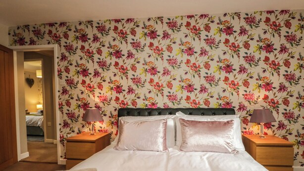A bedroom with a floral wallpaper, a bed with two pillows, two bedside tables with lamps, and a door leading to another room. - 1 Springfort Cottages (Penrith)