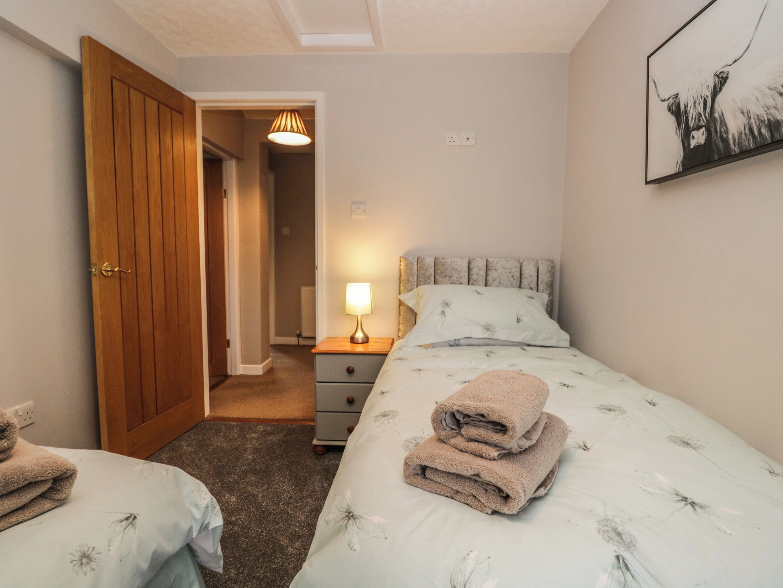 A bedroom with a wooden door, a bedside table with a lamp, a bed with floral bedding, and a framed picture on the wall.