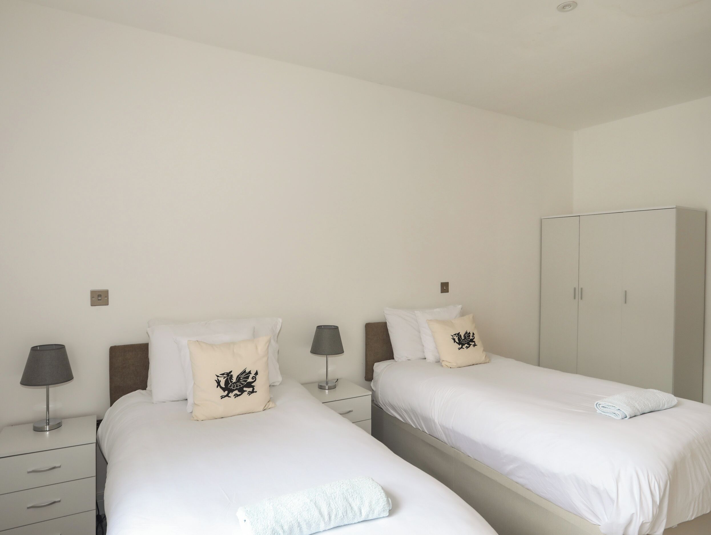 Quarto de hotel com duas camas de solteiro, cada uma com colcha branca e almofadas a combinar, mesa de cabeceira com abajur e guarda-roupa.