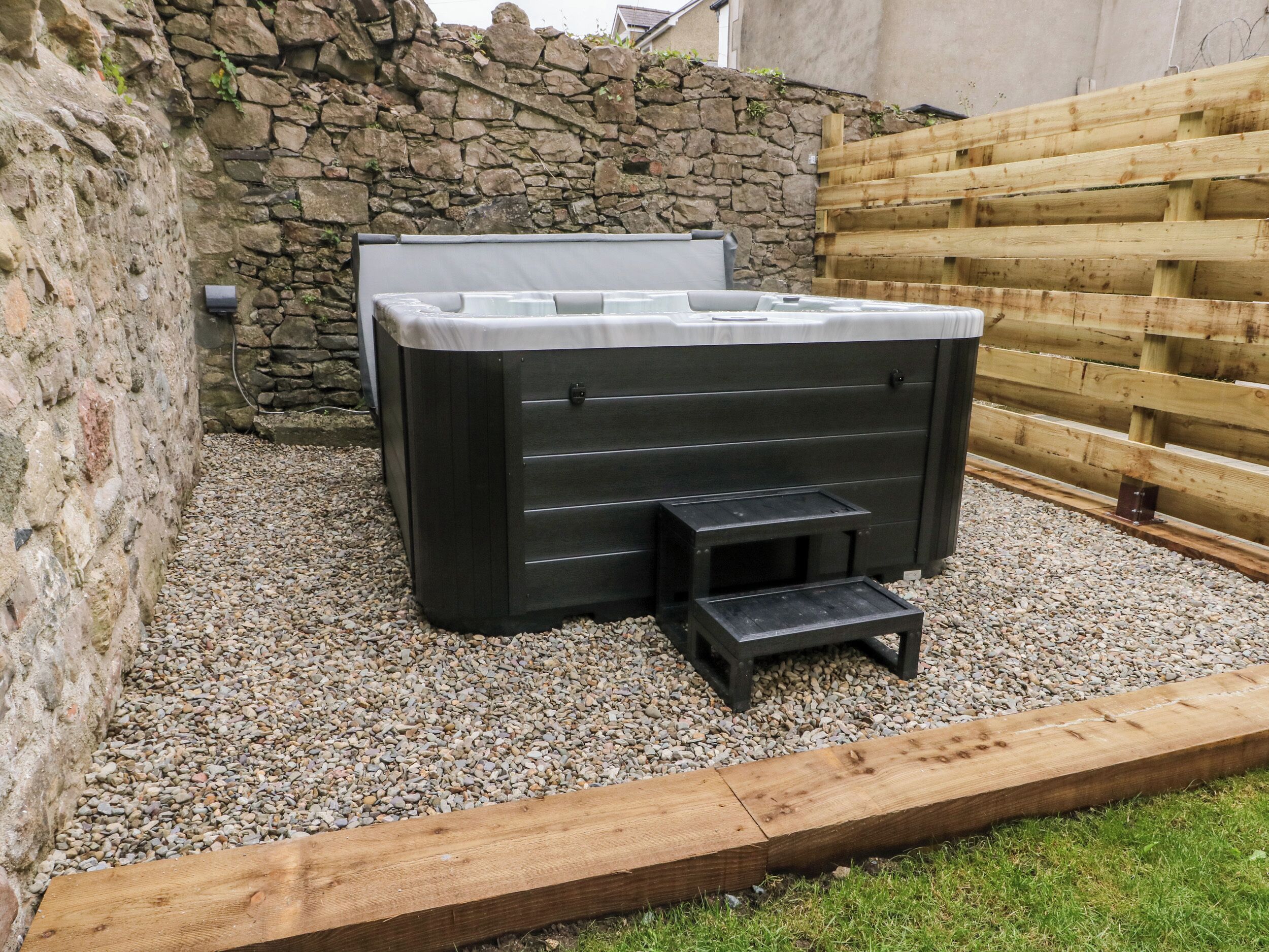 A hot tub in a garden area with a stone wall and wooden fence.
