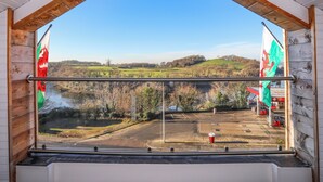 Vista desde una habitación con un ventanal grande que da a un paisaje con colinas, un río y un estacionamiento con coches y una gasolinera.