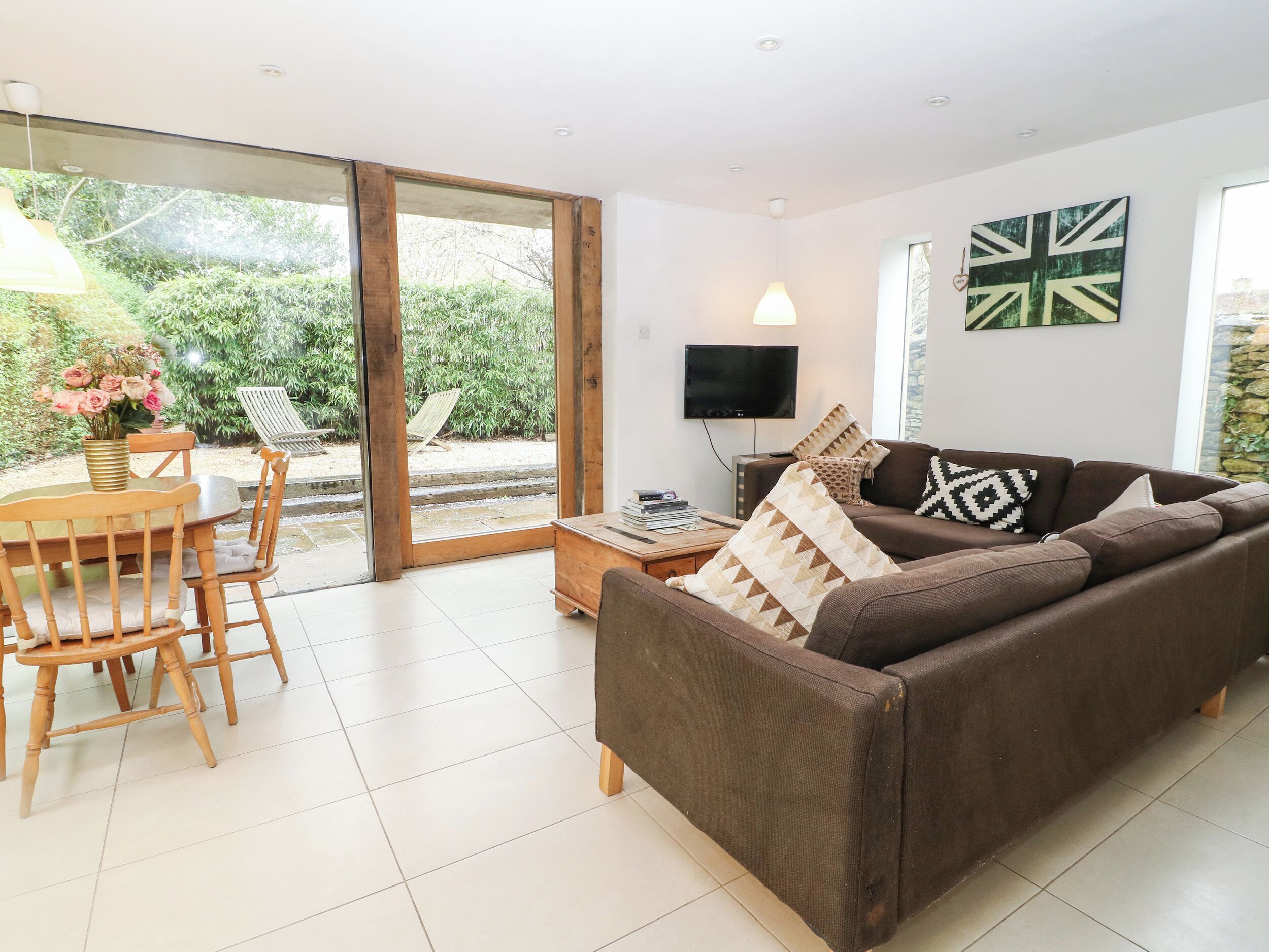 A living room with a brown sofa, a wooden dining table with chairs, a TV, and a large window overlooking a garden.