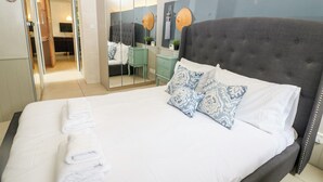 Un lit bien fait, avec du linge de lit blanc et des coussins à motifs, une tête de lit rembourrée de couleur grise et un grand miroir qui reflète la pièce.