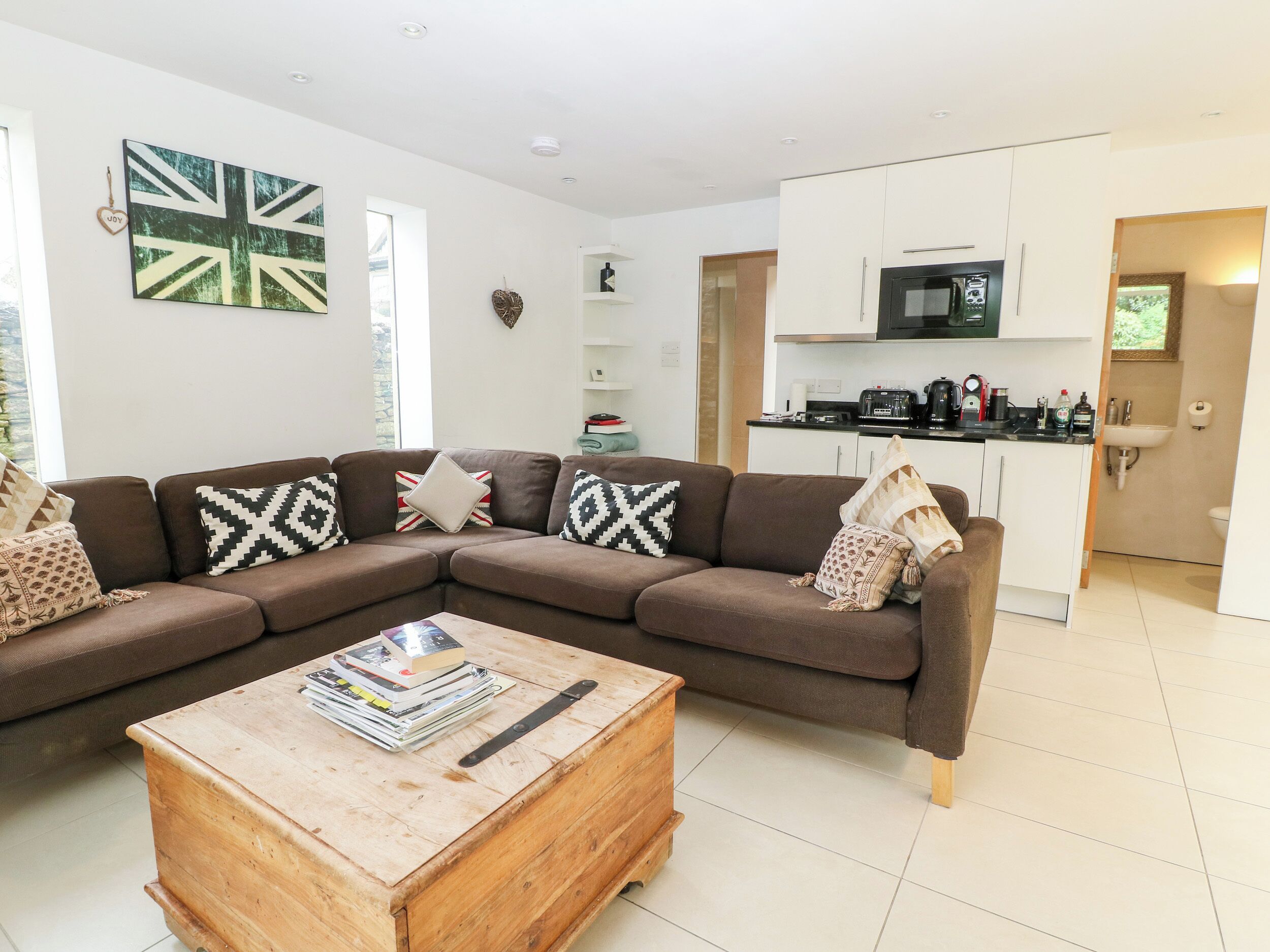 A modern living room with a brown sectional sofa, a wooden coffee table, and a kitchen area with white cabinets and a microwave.