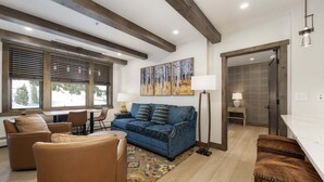 A modern living room with a blue sofa, brown leather chairs, and a wooden floor. There are large windows with blinds, a painting on the wall, and a lamp.