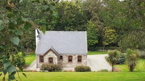 A stone house with a grey roof, surrounded by a well-maintained lawn and trees. - Cornode Cottage (Garrykennedy)