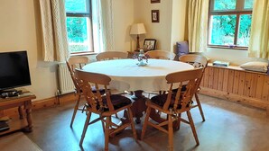 A dining room with a round table, six wooden chairs, a television, and two windows with curtains. - Cornode Cottage (Garrykennedy)