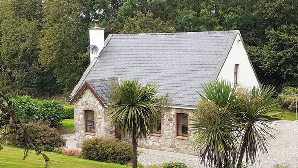 A stone house with a sloped roof, surrounded by lush greenery and a well-maintained lawn. - Cornode Cottage (Garrykennedy)