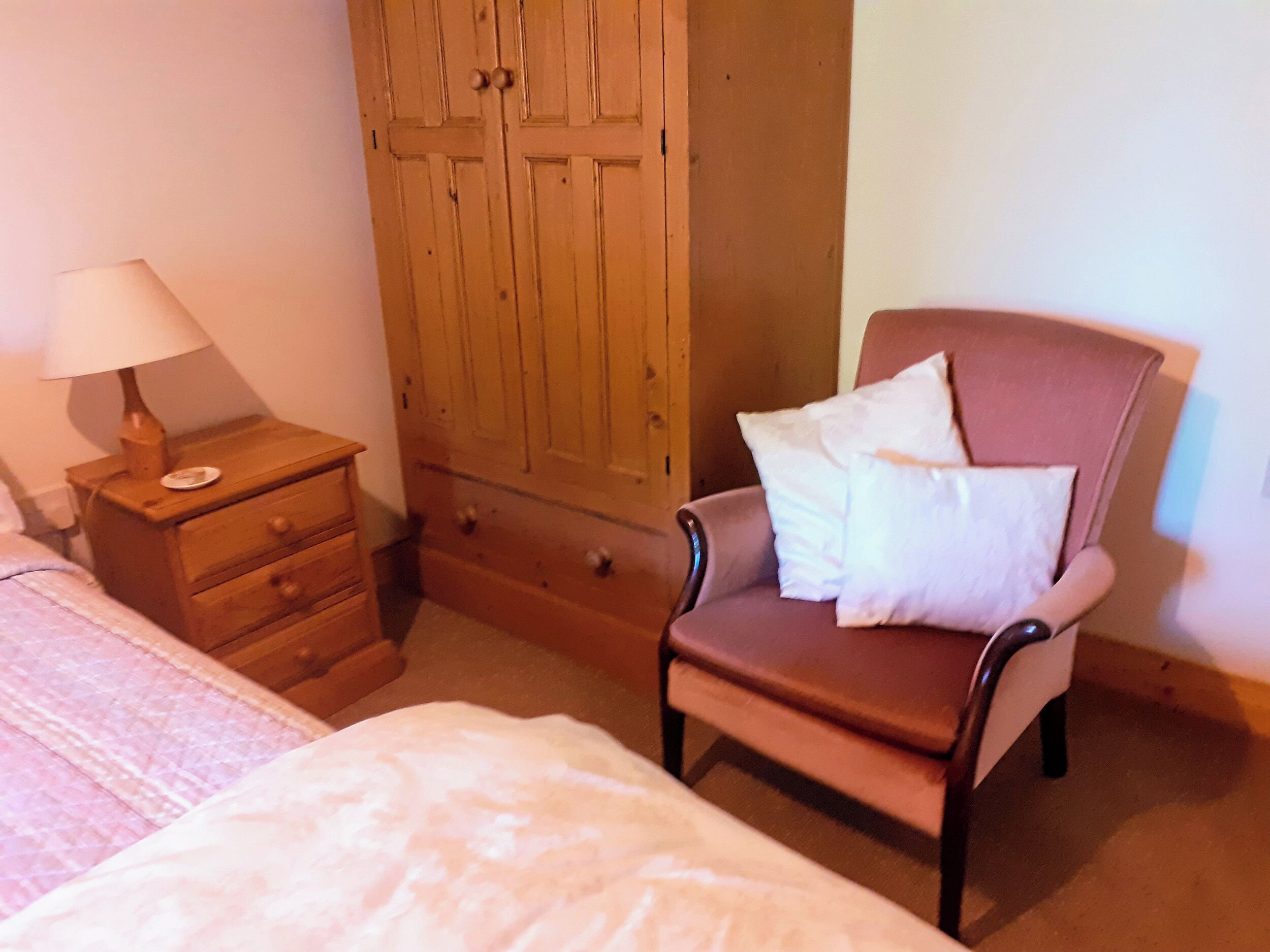 Un dormitorio con cama, mesita de noche con lámpara, armario de madera y una silla con cojines.