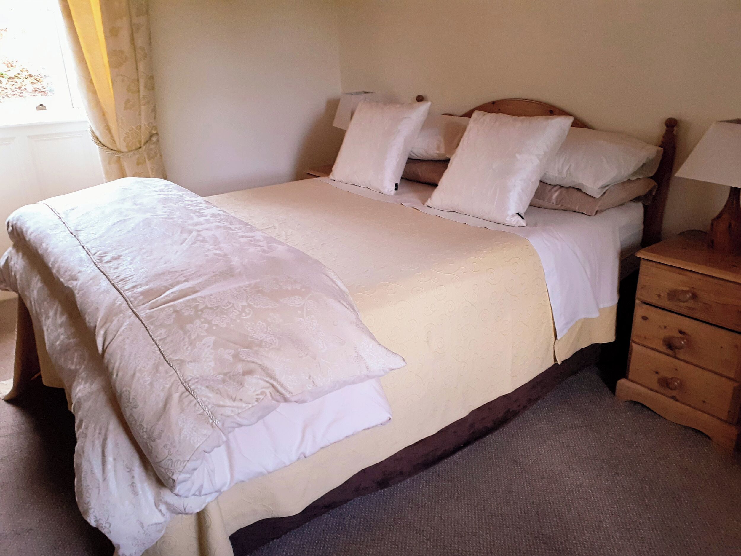 Una cama bien hecha con edredón blanco y almohadas, una mesita de noche de madera con cajones y una ventana con cortinas.
