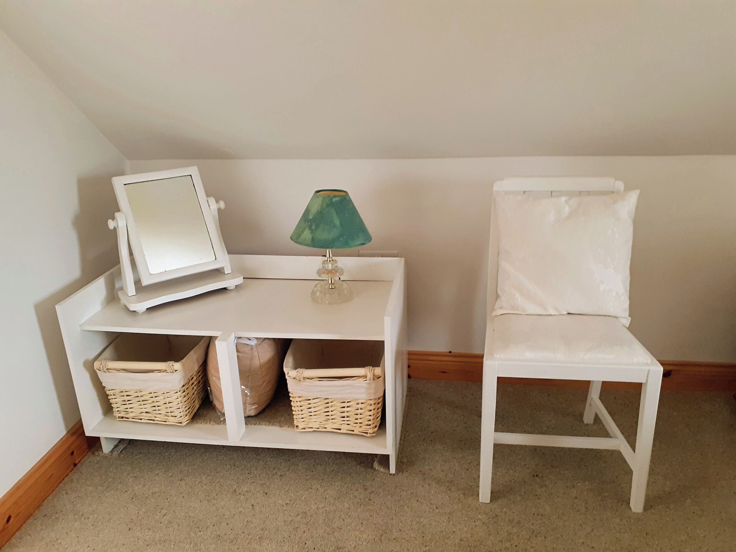 A small, white, corner room with a white desk, a white chair, and two wicker baskets on the floor.