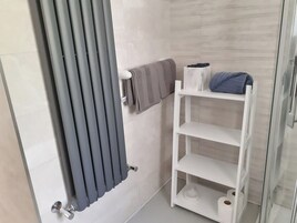 Un baño con un radiador blanco, un perchero con toalla, un estante blanco con artículos de aseo y un box de ducha de vidrio.