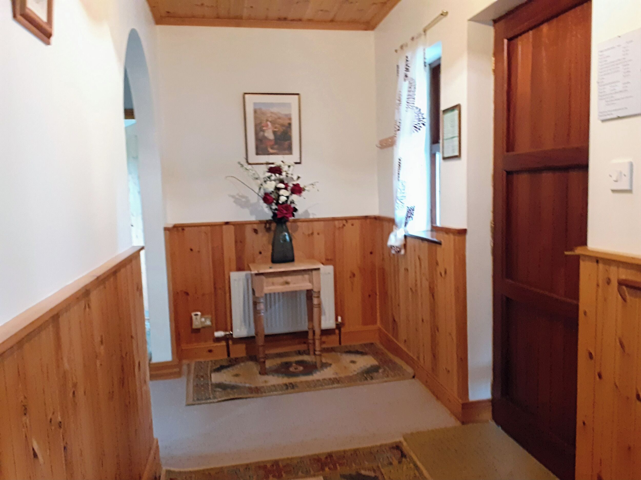 Ein Korridor mit Holzwandverkleidung, ein kleiner Tisch mit Blumen in einer Vase, ein gerahmtes Bild an der Wand und eine Tür mit Fenster.