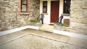 A stone building with a wooden door and windows, a porch with chairs, and a gravel area. - Fraoch Cottage (Garrykennedy)