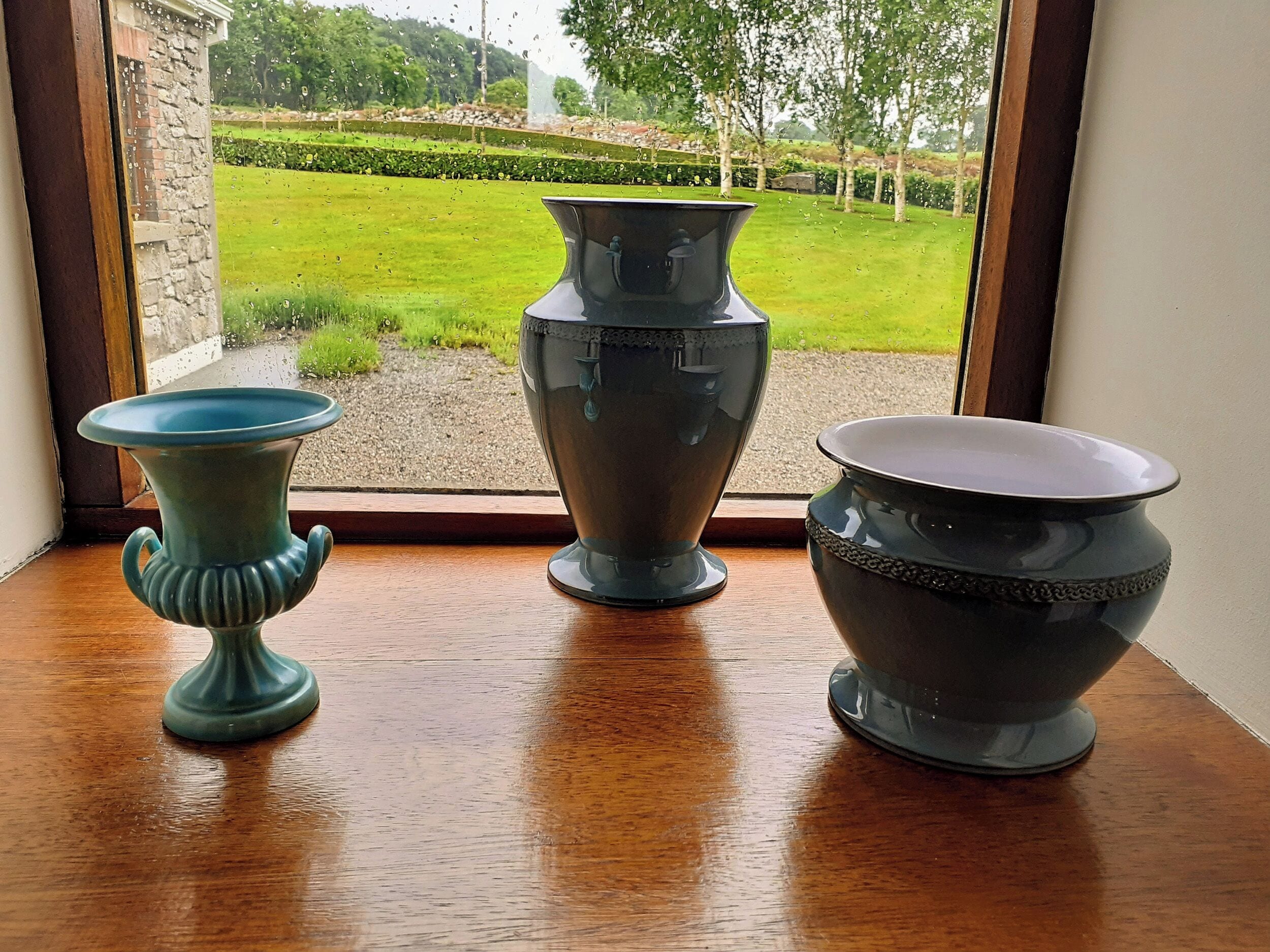 Tres jarrones sobre una mesa de madera junto a una ventana con vistas a un paisaje verde.