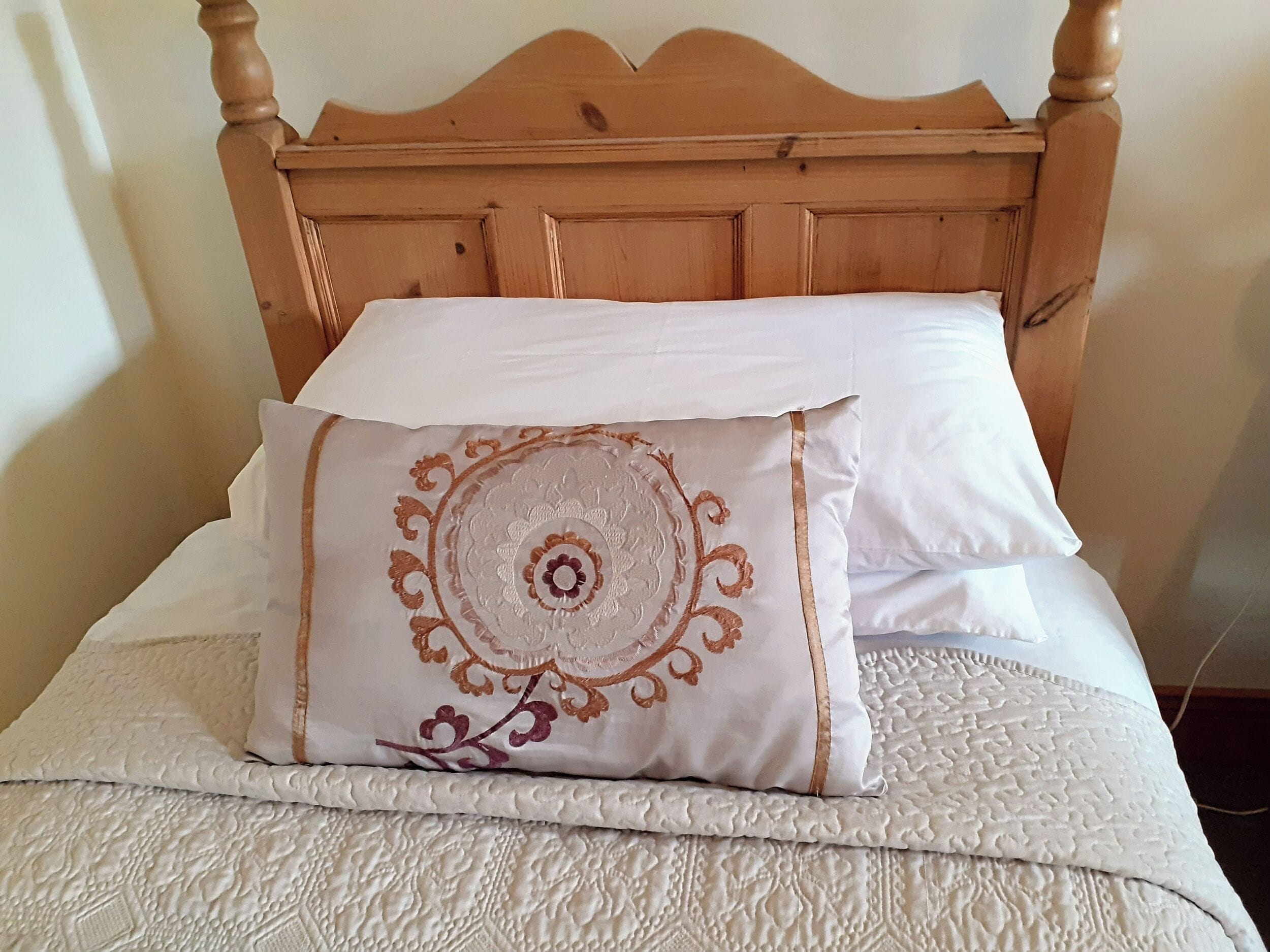 Un lit avec une tête de lit en bois, une couvre-lit blanche matelassée et un coussin décoratif orné d’un motif circulaire.