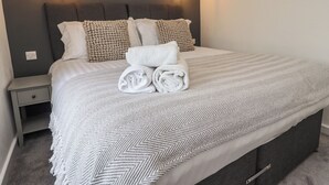 A neatly made bed with rolled towels, a grey headboard, and two wall-mounted light fixtures.