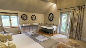 A bedroom with a large bed, two bedside tables, a sitting area with a chair, a bathroom with a bathtub, and a view of the outdoors. - The Stables (Ledbury)