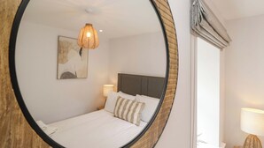 A bedroom with a round mirror, a bed with striped pillows, a bedside lamp, and a wall-mounted artwork. - Riverside Cottage No 5 (Keswick)