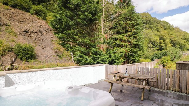 A hot tub with a wooden deck and a wooden fence, surrounded by a hillside with dense trees. - Riverside Cottage No 5 (Keswick)