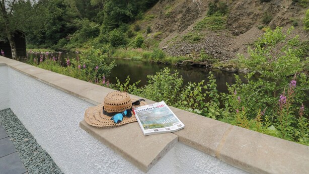 A stone bench with a straw hat and sunglasses, and a magazine on it, overlooking a river with greenery and trees. - Riverside Cottage No 4 (Keswick)