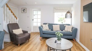 A living room with a grey sofa, a brown armchair, and a round coffee table with a plant. A staircase with white railings is visible in the background. - Riverside Cottage No 4 (Keswick)