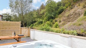 A hot tub with a wooden table and a bottle, surrounded by a white wall and a wooden fence, with a forested hillside in the background. - Riverside Cottage No 4 (Keswick)