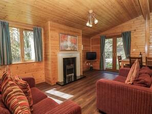 A wooden cabin interior with a fireplace, sofa, television, and dining area. - Elm Lodge (Keswick)