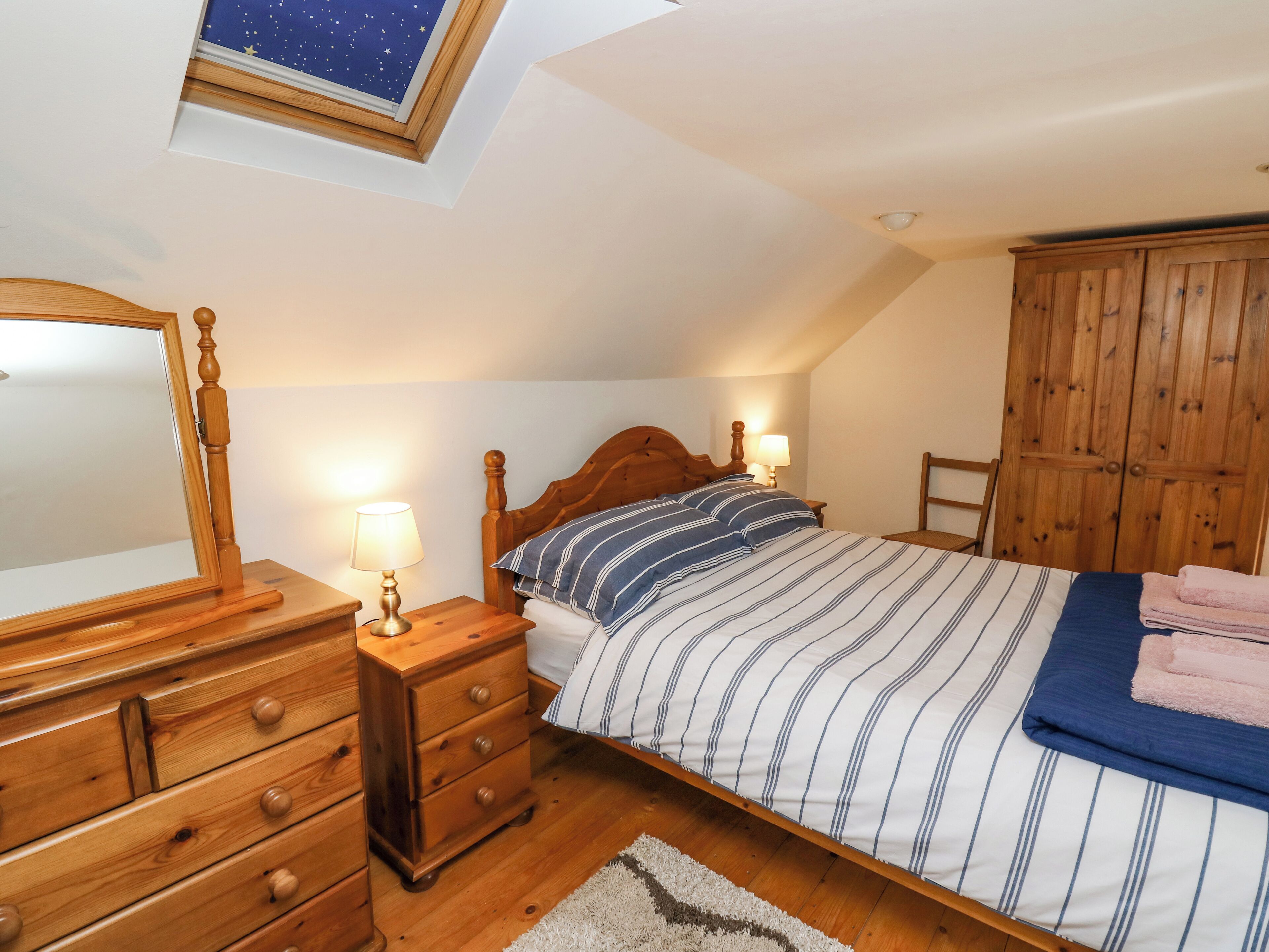 A bedroom with a wooden bed, a nightstand with a lamp, a dresser, a wooden wardrobe, and a skylight above the bed.