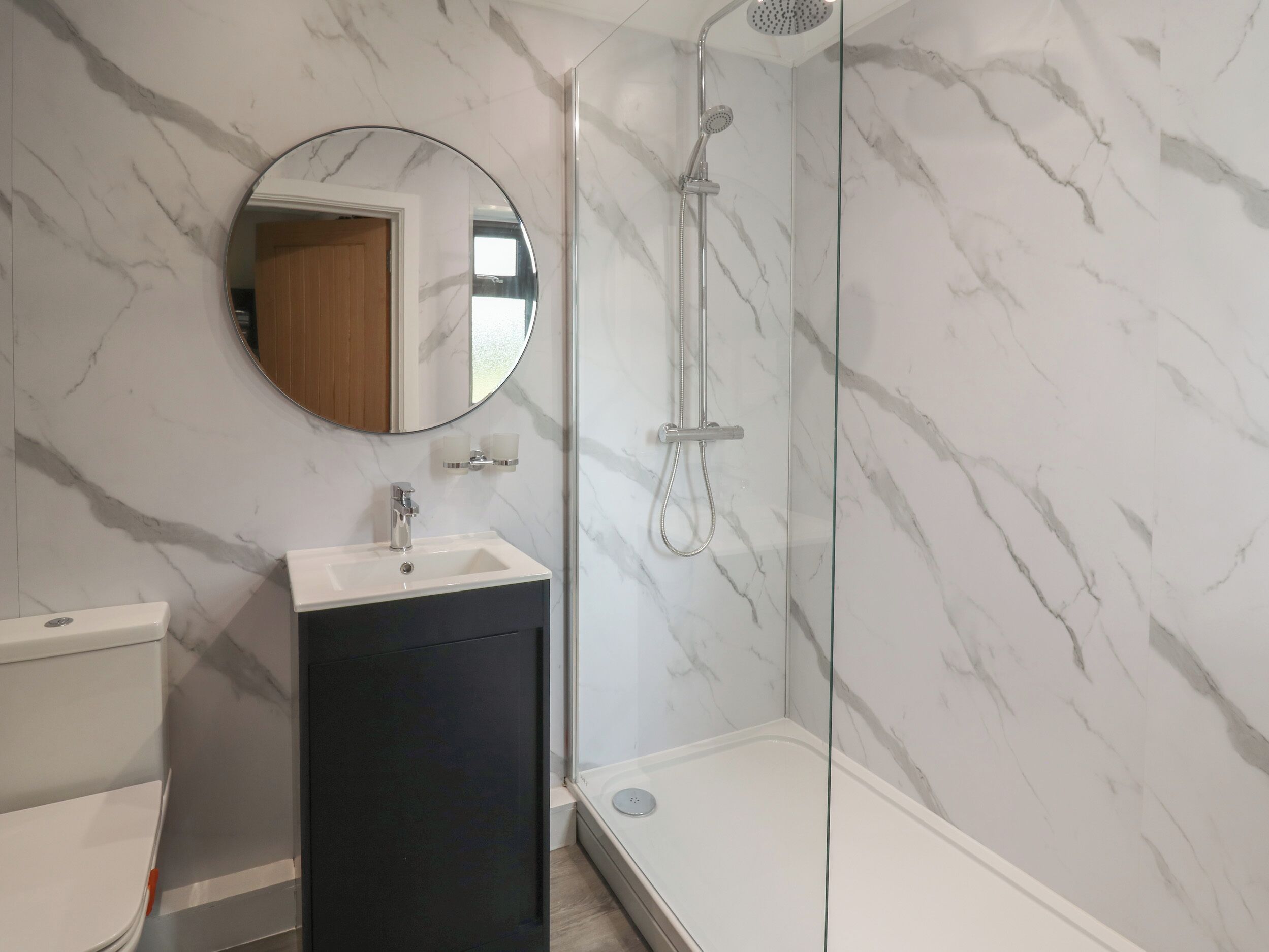 A bathroom with a marble wall, a round mirror, a sink, a toilet, and a glass-enclosed shower.