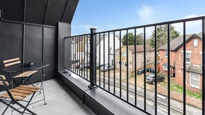 Un balcó amb barana de metall negre, taula petita amb una cadira i vista a un carrer residencial.
