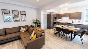 A modern living room with a brown sofa, a dining area with a wooden table and chairs, and a kitchen with dark wooden cabinets. - Briery House (Keswick)