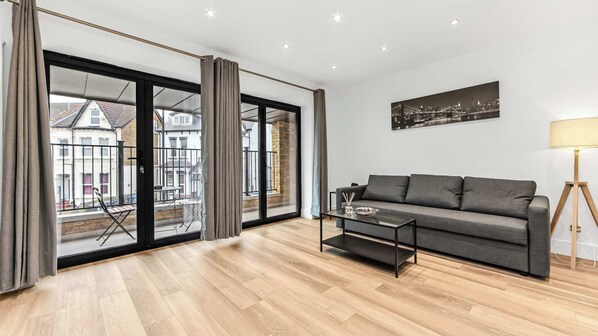 Ein modernes Wohnzimmer mit einem grauen Sofa, einer Glastür, die auf einen Balkon im Freien führt, und Blick auf die Stadt.
