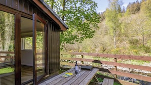 Eine Holzterrasse mit Glastür zu einem Raum, einem Picknicktisch und Blick auf Fluss und Wald.
