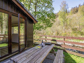 Une terrasse en bois avec une porte vitrée donnant sur une pièce, une table de pique-nique et une vue sur une rivière et une forêt.