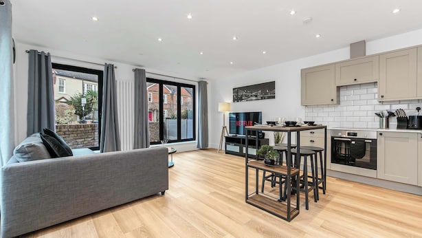 Un apartamento moderno con un sofá gris, una mesa de comedor con sillas, una cocina con armarios blancos y un ventanal con cortinas.